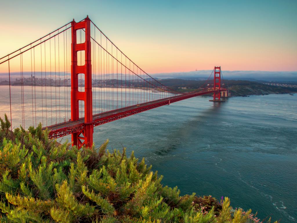 A scenic view of Golden Gate Bridge from Battery Spencer, Golden Gate National Recreation Area with a beautiful hue of orange and yellow sunrise in a clear blue sky.