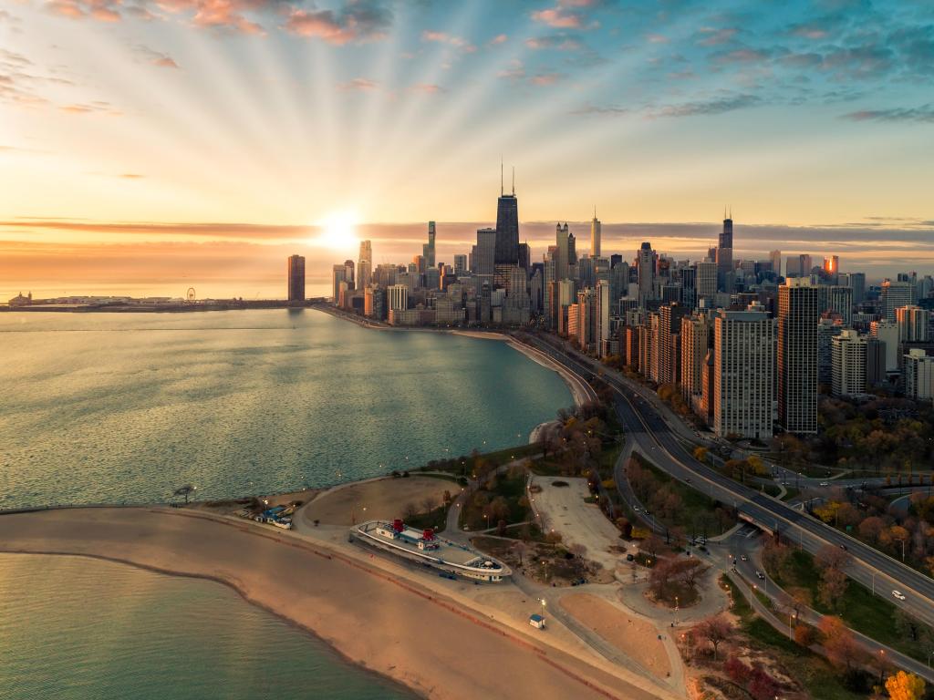 High rise buildings run along the lake shore with a sandy beach in the foreground and the rising sun illuminating the city