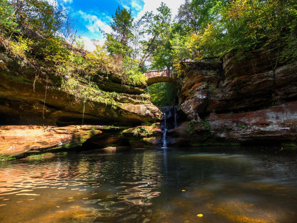 Hocking Hills State Park, near Columbus with a beautiful waterfall surrounded by rocks and a stone bridge in the center, surrounded by trees.