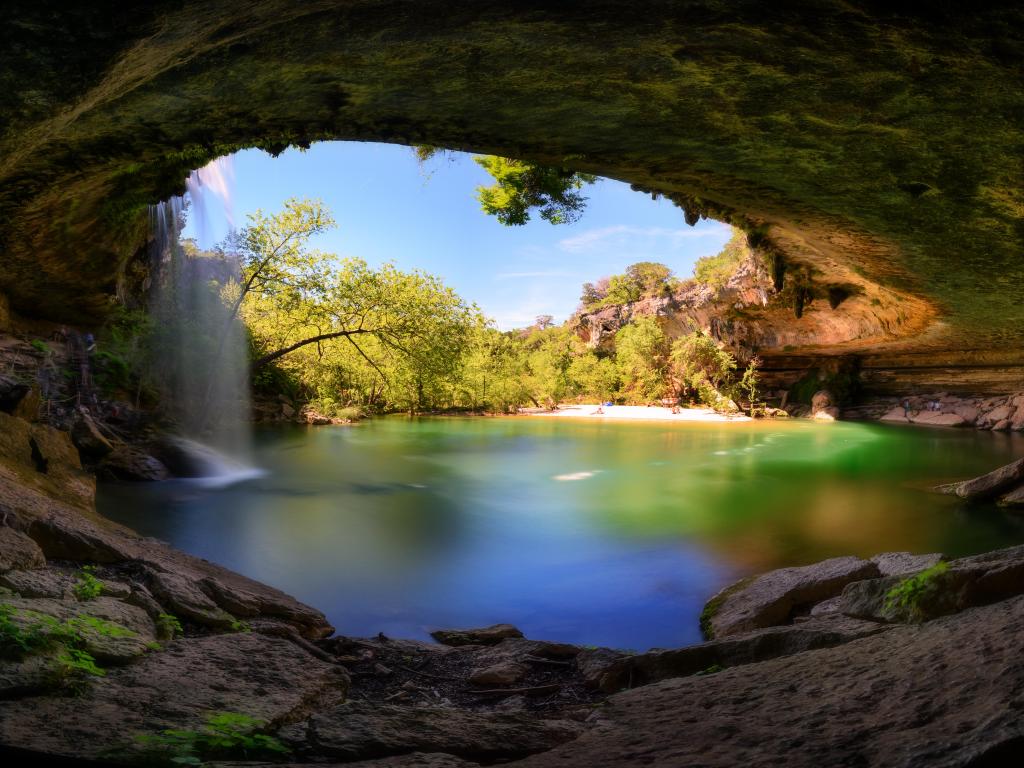 Hamilton Pool, Texas, USA with a view of a waterfall, turquoise water, beach and trees with rocks framing the photo.