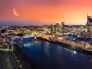 Nashville, Tennessee with the city in the background and the river in the foreground at night with the moon visible in a red sky.