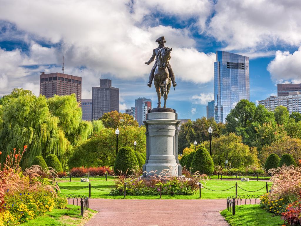 Boston, Massachusetts with the George Washington Monument in the foreground surrounded by the green park, with flowers and the city skyline in the distance on a cloudy but sunny day.