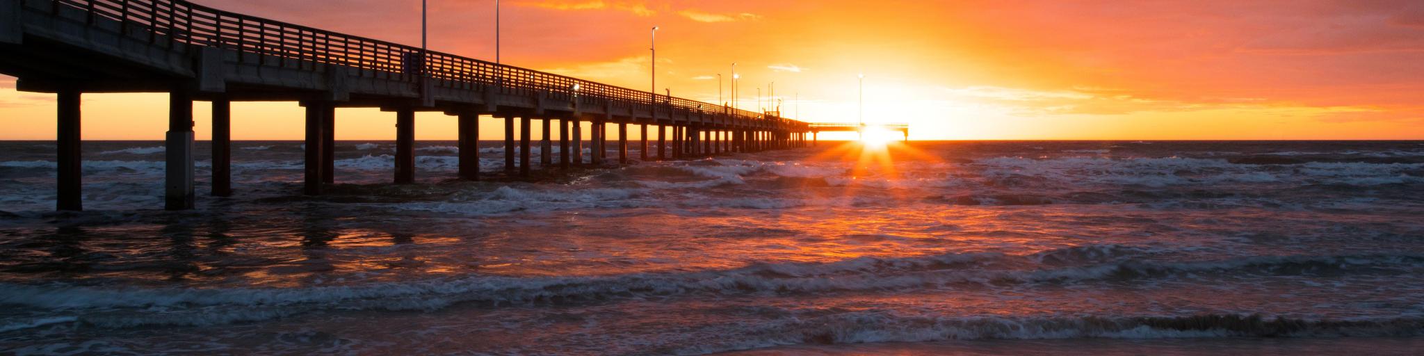 Corpus Christi, Texas, USA with the Bob Hall Pier on Padre Island at sunset with the sun low against the sea and beach in the foreground.