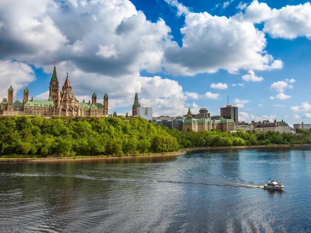 Parliament Hill, Ottawa, Ontario, Canada taken on a sunny day with a boat in the water, trees lining the shore and the buildings in the background.