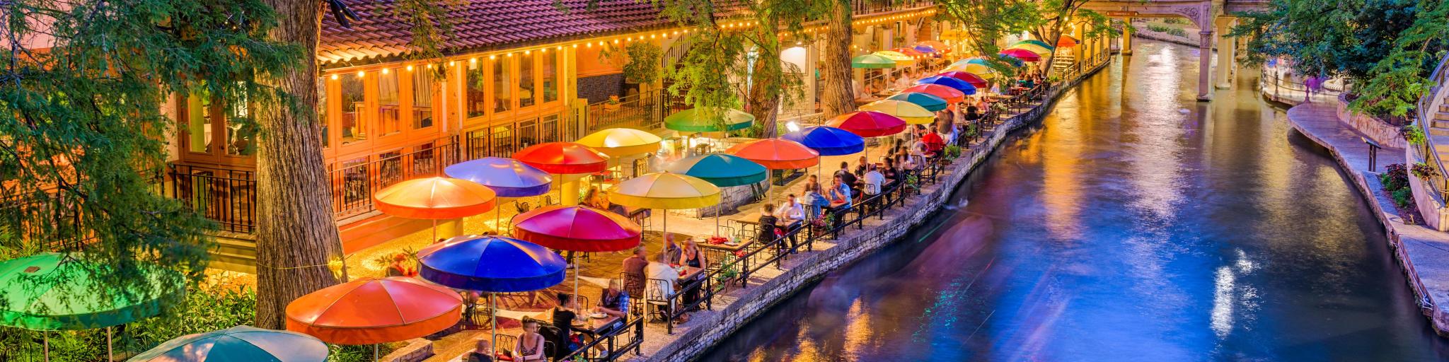 San Antonio, Texas, USA cityscape at the River Walk as the evening falls. There are colorful umbrellas on the riverside, with tables.