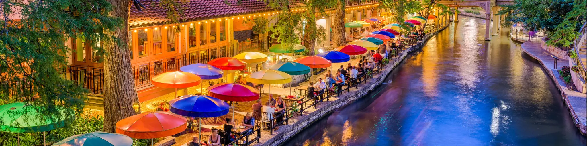 San Antonio, Texas, USA cityscape at the River Walk as the evening falls. There are colorful umbrellas on the riverside, with tables.