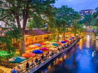 San Antonio, Texas, USA cityscape at the River Walk as the evening falls. There are colorful umbrellas on the riverside, with tables.