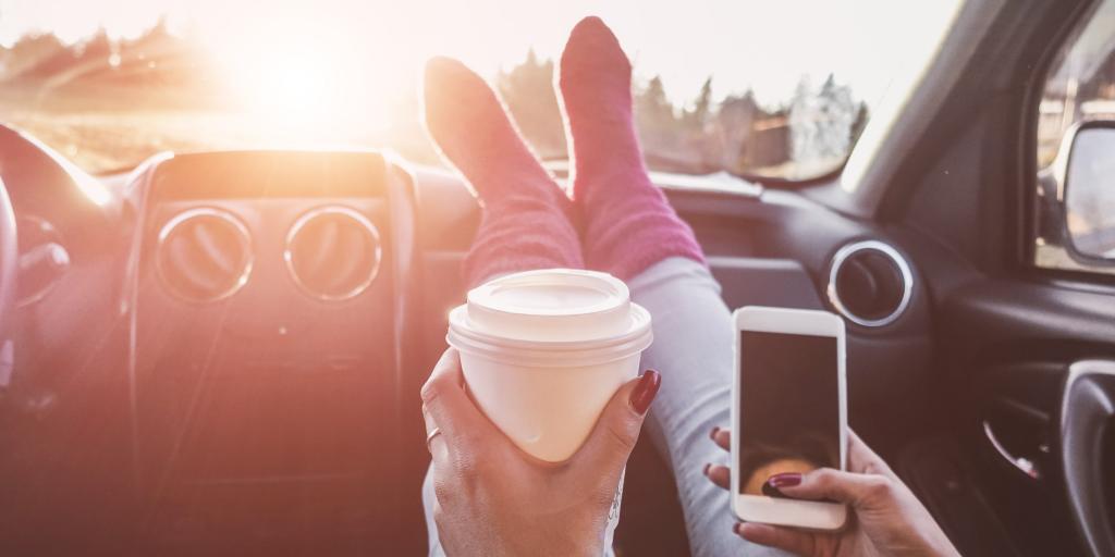 A person with their feet on a car dashboard holding a coffee in one hand and their phone in the other