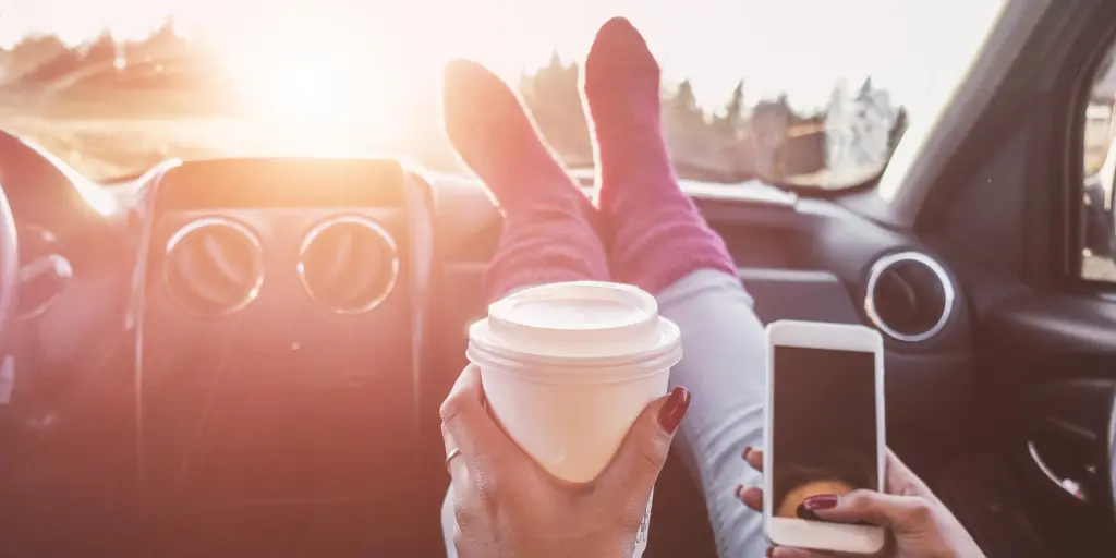 A person with their feet on a car dashboard holding a coffee in one hand and their phone in the other
