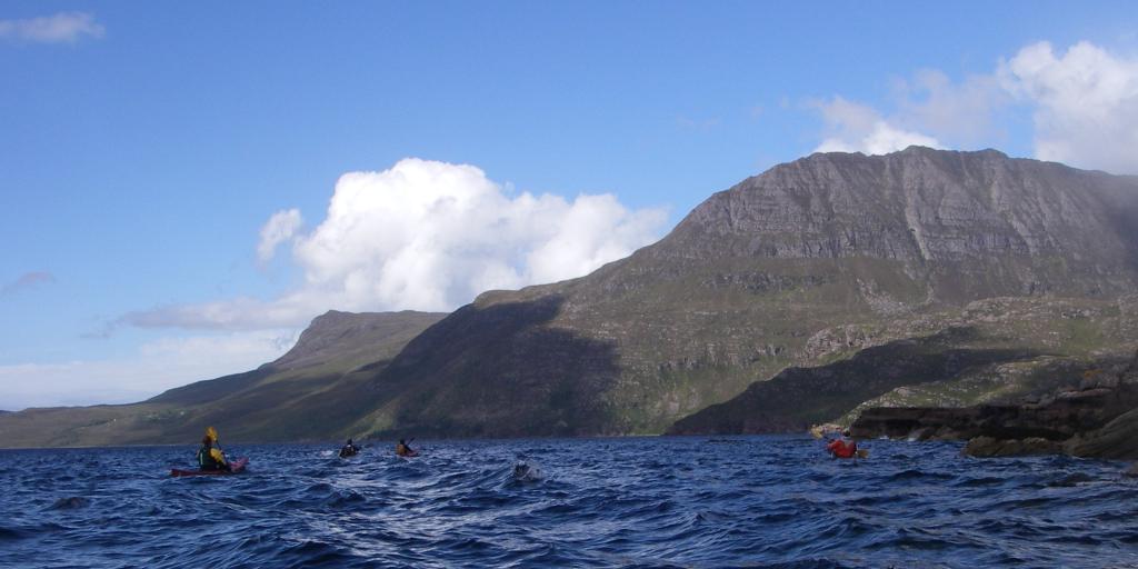 The tip of a kayak in the water, with other kayaks and mountains in the background