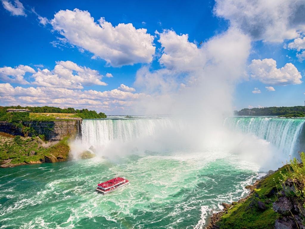 Niagara Falls, Canada taken on a sunny day with clouds and a red boat in the foreground and the stunning waterfall creating a spray. 