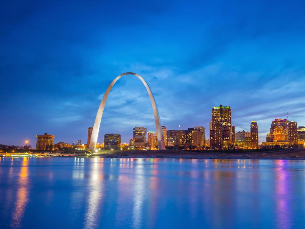 At twilight, a large arch monument is illuminated on a riverbank with downtown buildings behind
