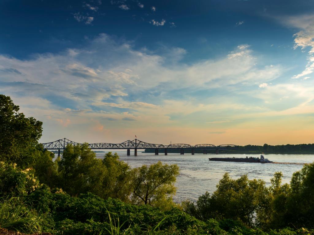 Vicksburg, Mississippi, USA with a boat in the Mississippi River near the Vicksburg Bridge at sunset, trees in the foreground.