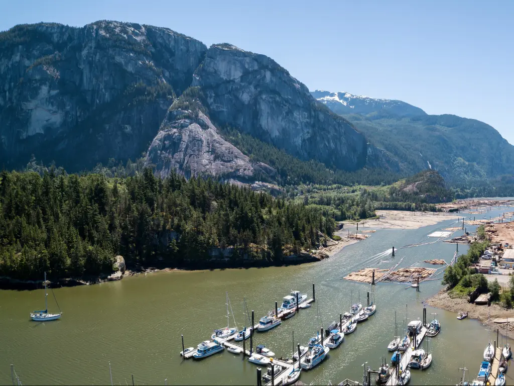 Squamish, British Columbia, Canada taken as a panoramic aerial view of the town, surrounded by boats and mountains with a dense forest on a clear sunny day.