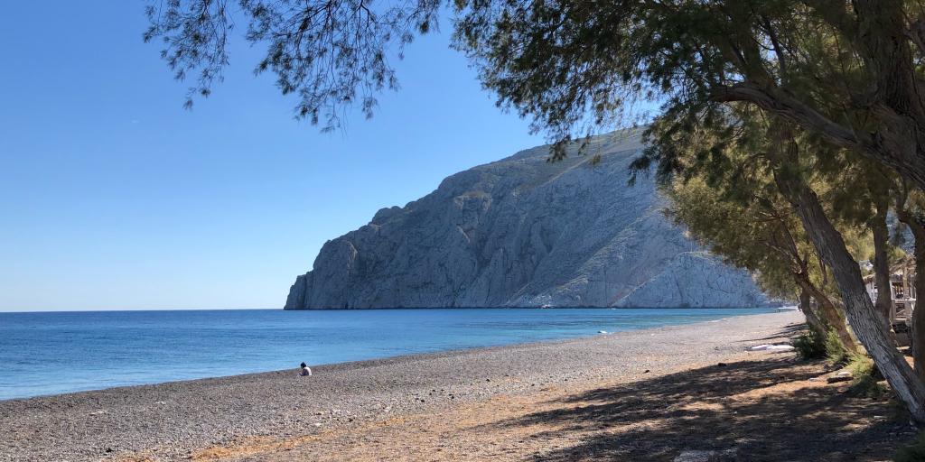 A tree overhangs the empty Kamari Beach on Santorini, with dramatic cliffs in the background