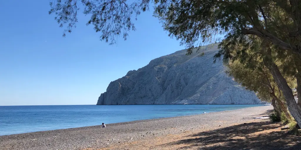 A tree overhangs the empty Kamari Beach on Santorini, with dramatic cliffs in the background