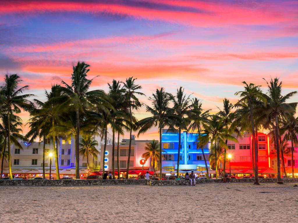 Miami Beach, Florida on Ocean Drive at sunset, with a beach in the foreground and palm trees before a row of buildings lit up in fluorescent colours.