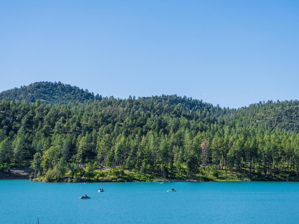 View of blue waters of Mezcalero with boaters dotted in the water, surrounded by lush tall trees and hills of Lincoln National Forest