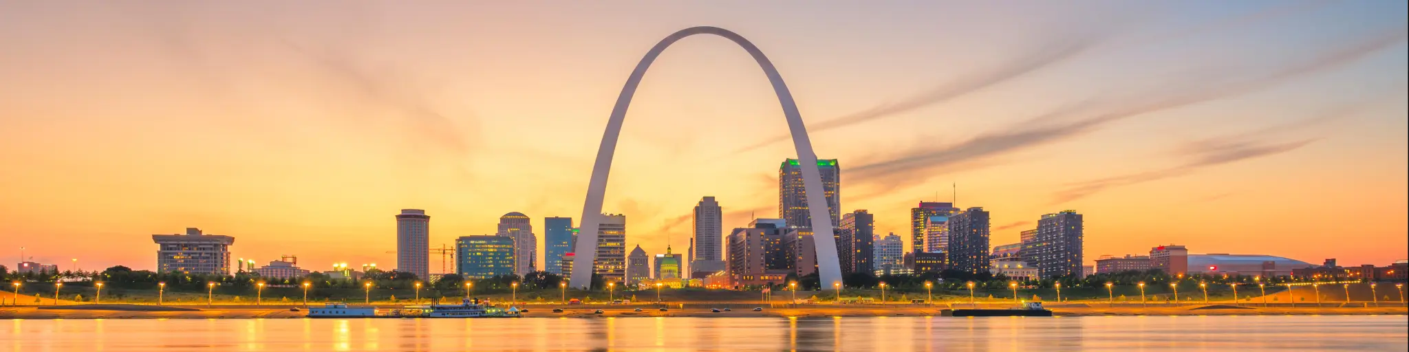 A sunset shot of the cityscape of St Louis with the famous arch in the foreground, overlooking water