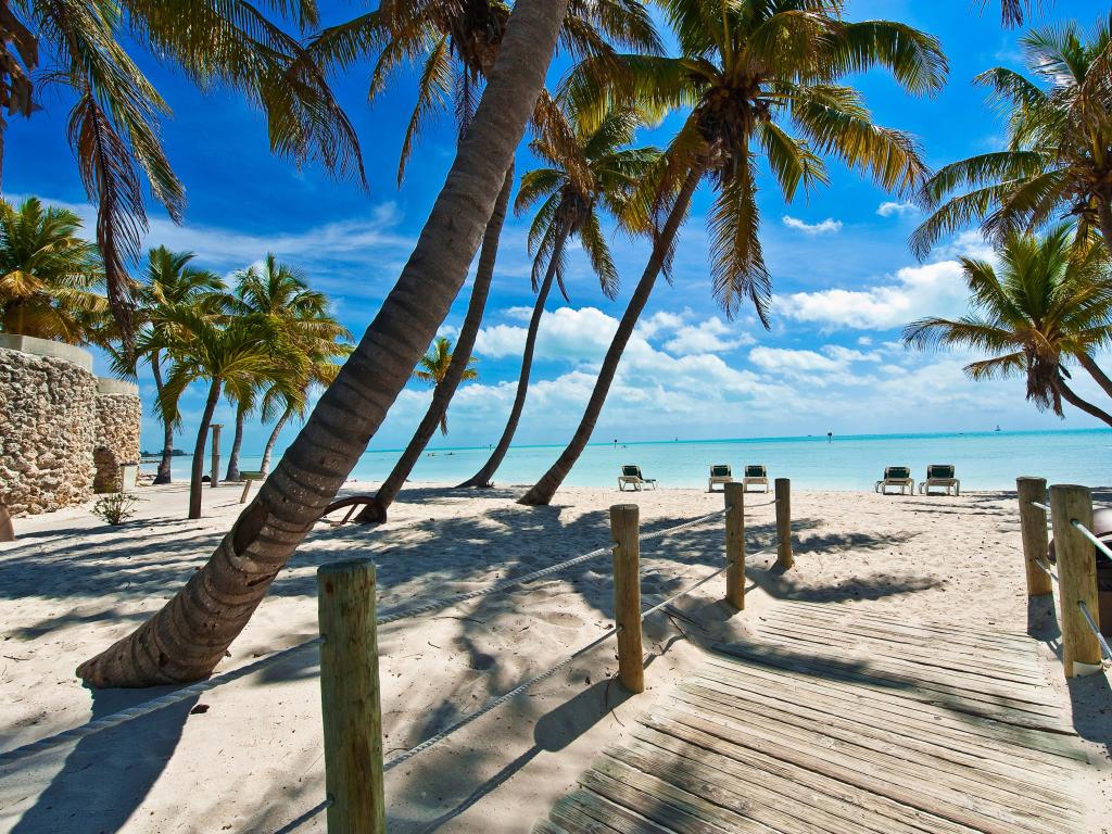 Key West, Florida with a footbridge in the foreground leading to the beach with palm trees both sides and the turquoise water in the distance on a sunny day.