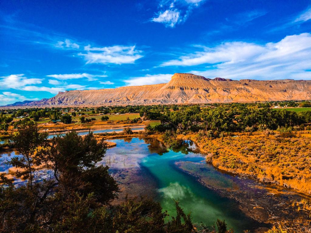 Grand Mesa, Uncompahgre And Gunnison National Forests, USA with the Colorado River in the foreground and surrounded by orange land, trees and mountain formations in the background on a sunny clear day.