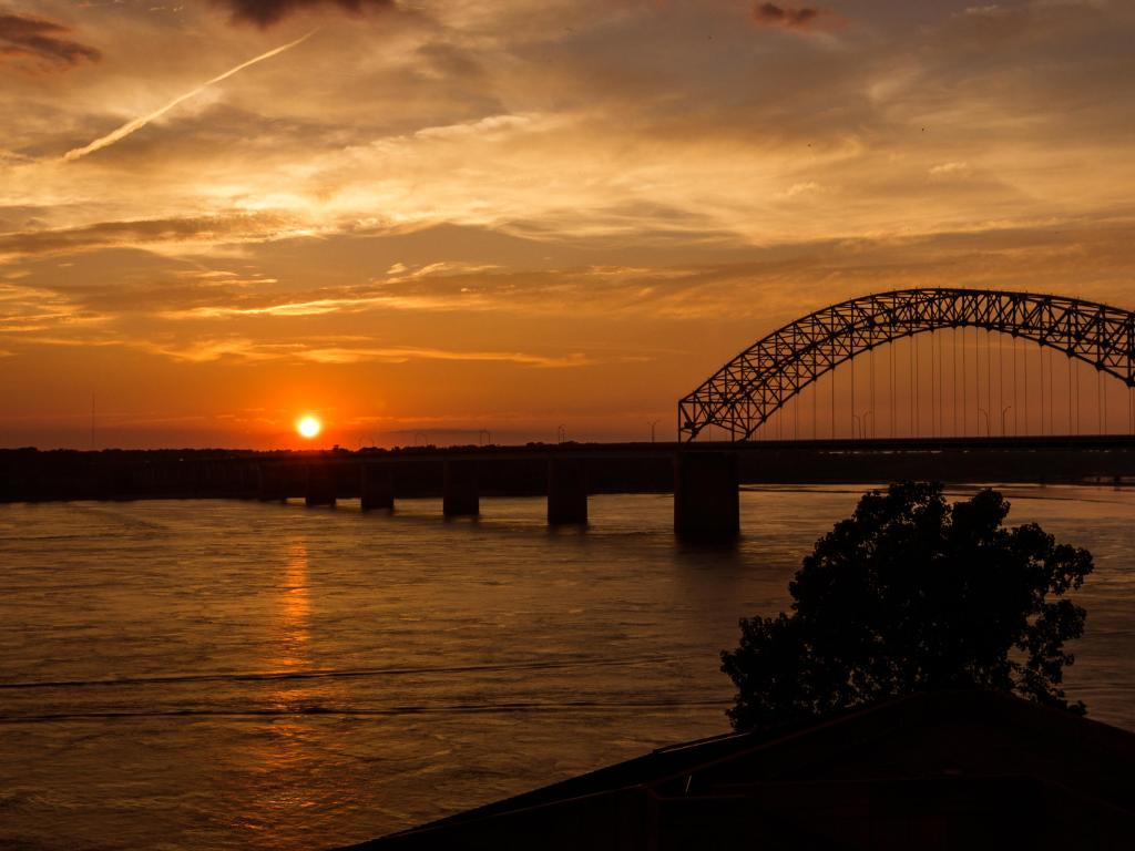 Long bridge over wide river with banks shown in silhouette and deep sunset light reflected on the water
