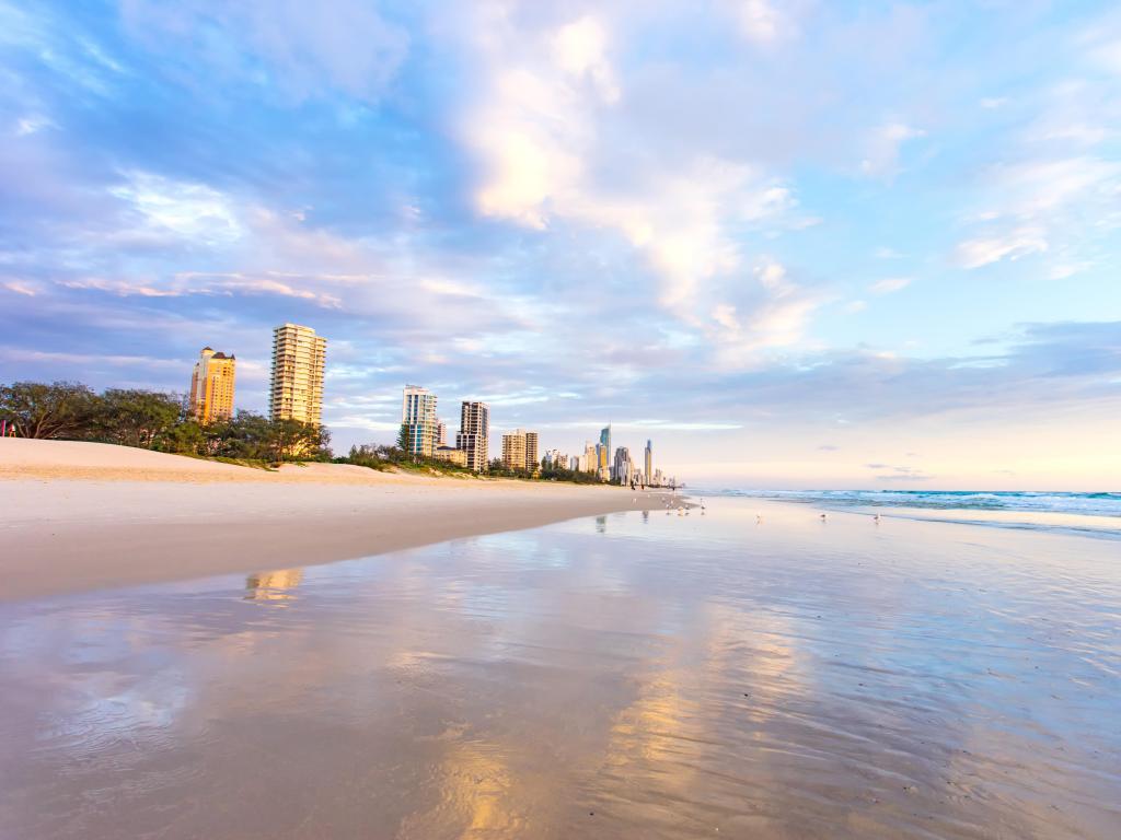 Surfers Paradise, Australia at sunrise with reflections of Queensland's Gold Coast and city buildings in the wet sand in the foreground.