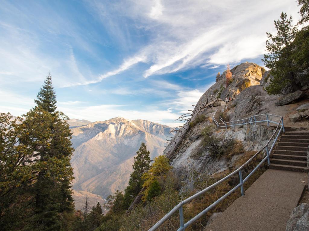 Sequoia National Park, California at sunset on an autumn evening at Moro Rock in with steps alongside the mountain and trees in the foreground, plus more mountains in the distance. 