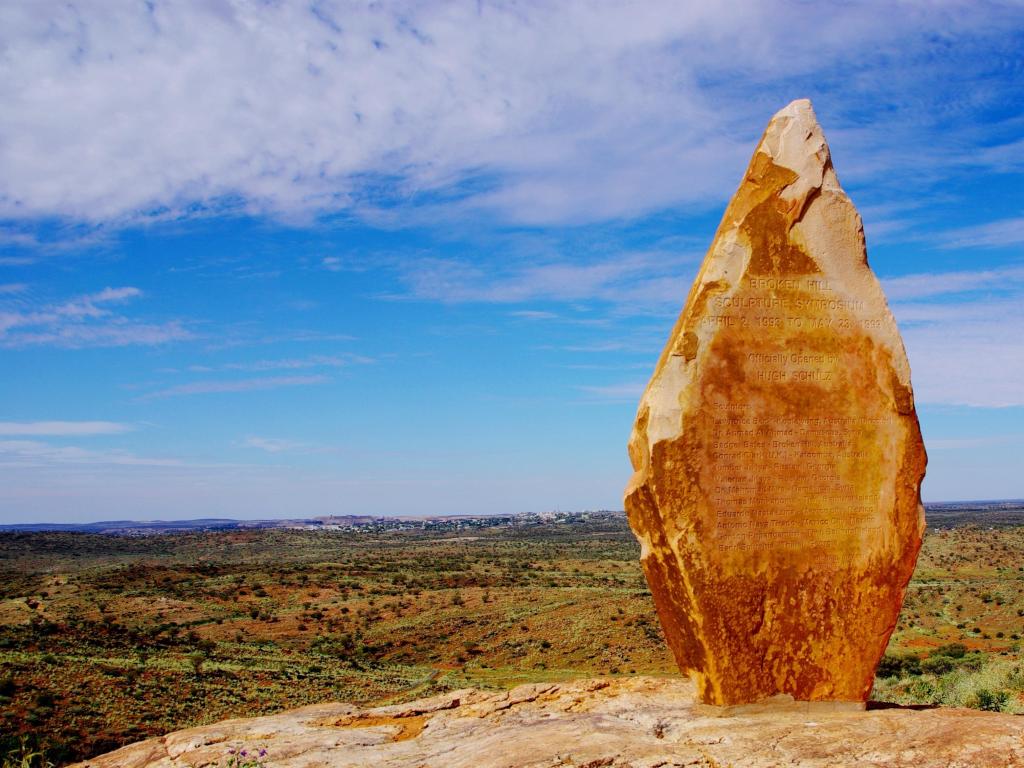 A sculpture made from rock placed on a hilltop overlooking sandy outback landscape with scrubby green vegetation