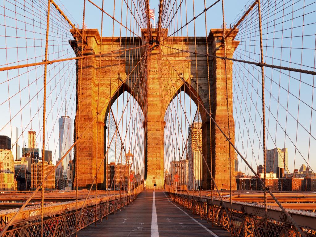 Brick arches across pedestrian walkway over bridge illuminated in warm light, with city skyline in the background, criss-crossed with bridge architecture