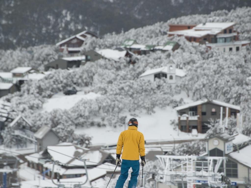 Male wearing bright coloured skiing clothes skiing on slope towards the village in the background all covered in snow