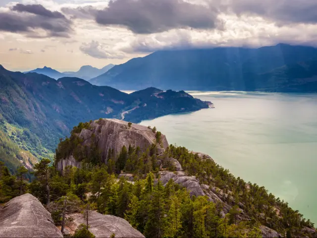 Howe Sound from the summit of Stawamus Chief, Squamish, British Columbia, Canada on a cloudy day with mountains in the distance and rocks and trees overlooking the lake.