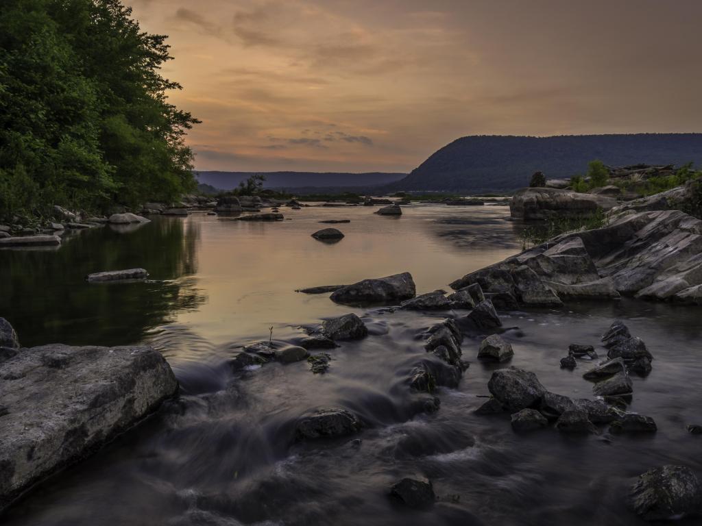 Calm river water appears silvery grey in sunset light, with tree-lined hillside in the background