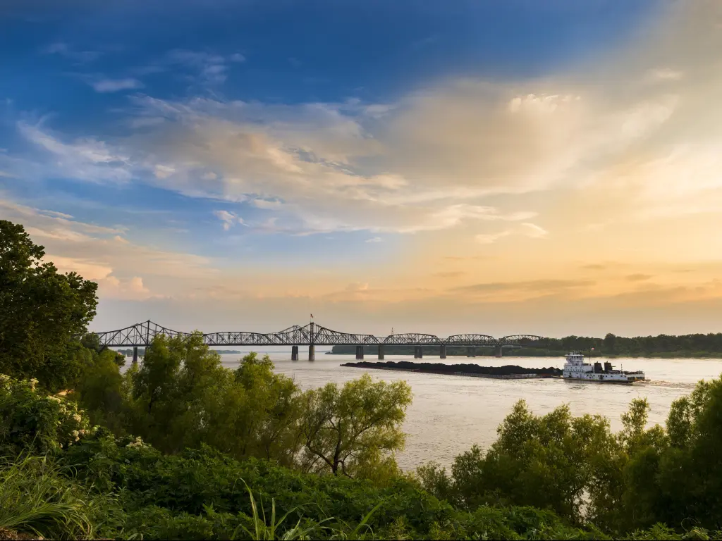 Vicksburg Bridge in Vicksburg, Mississippi with a boat in the Mississippi River, green foliage in the foreground and the bridge in the distance at sunset.