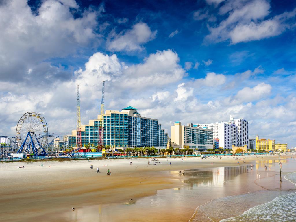 Daytona Beach, Florida, USA with the beachfront skyline, a gentle sea and large sandy beach in the foreground, rides and a blue sky in the background. 