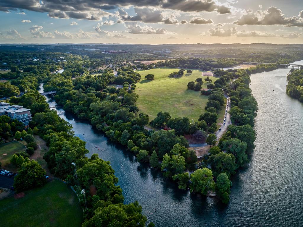 Zilker Park in Austin, aerial photo taken during a sunset with some clouds in the sky. The image depicts the water shores dotted with trees.
