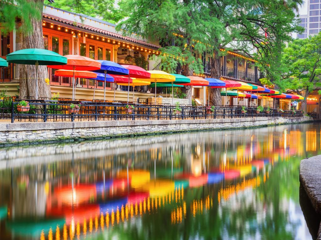 San Antonio, Texas, USA with the cityscape at the River Walk reflected in the waters edge, colorful umbrellas along the waterfront and trees lining the shore.