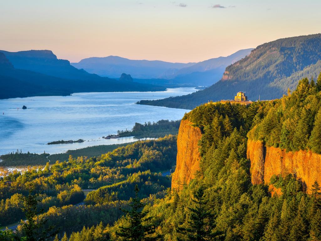 Wide river runs through a forested valley with sloping hillsides and sheer orange stone rock cliffs