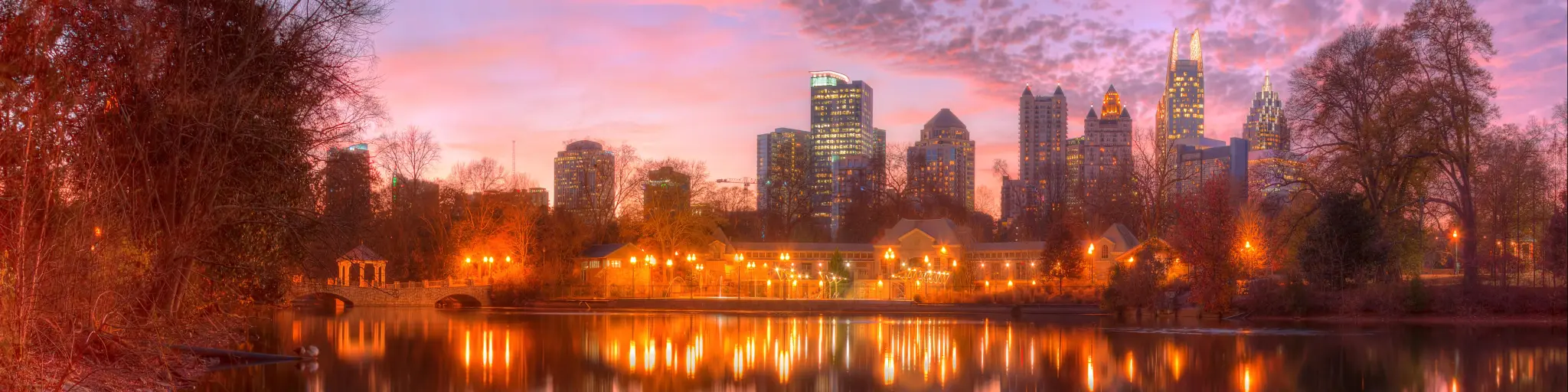 Panoramic view of Lake Clara Meer, with Piedmont Park Aquatic Center illuminated at dusk and Midtown Atlanta skyline in the background
