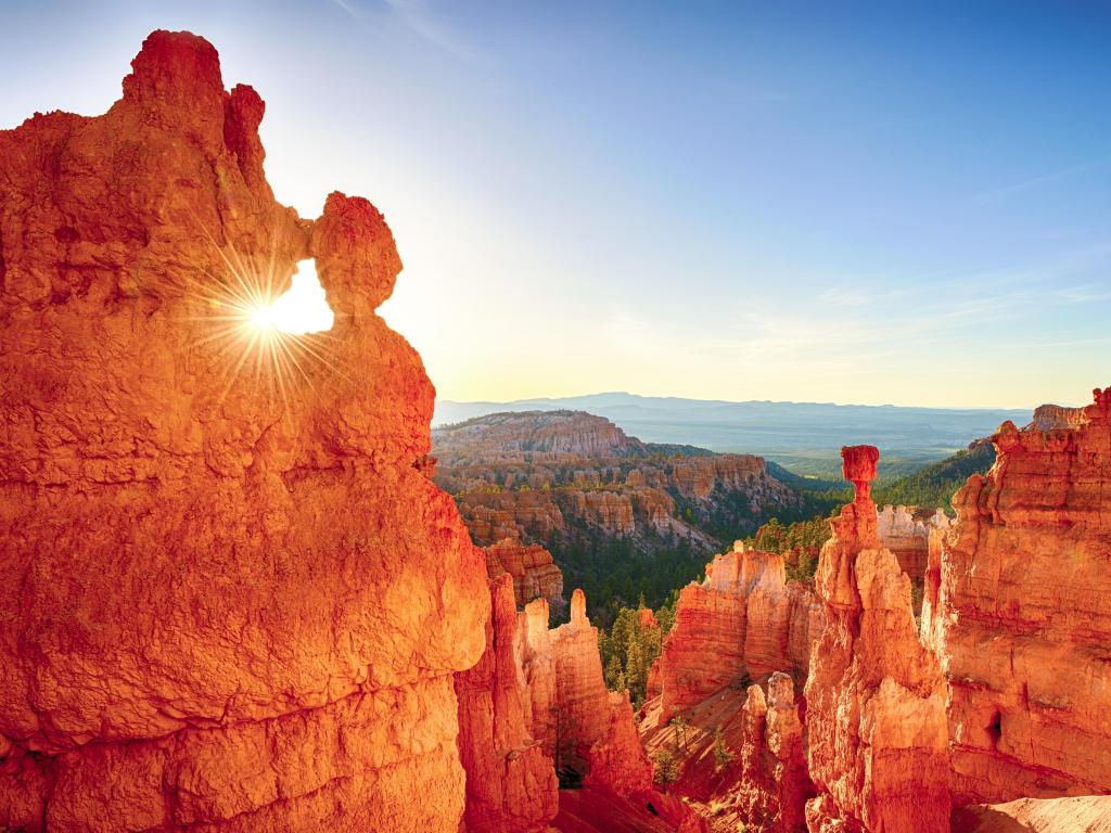 Sun shines through a natural window of red rock overlooking a deep red rock and green forest canyon