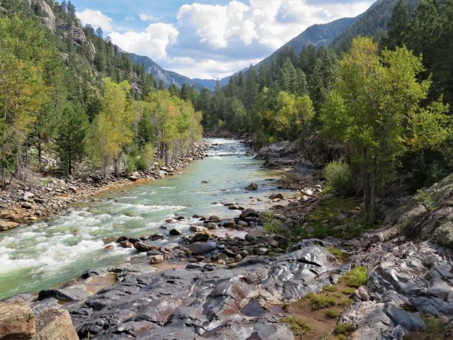 The winding Animas River running through Durango with trees on either bank and mountains in the distance. Water babbling over rocks.