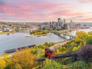 Pittsburgh, Pennsylvania at sunset with a view of a red mountain cart leading down to the city in the horizon and the river running through downtown.