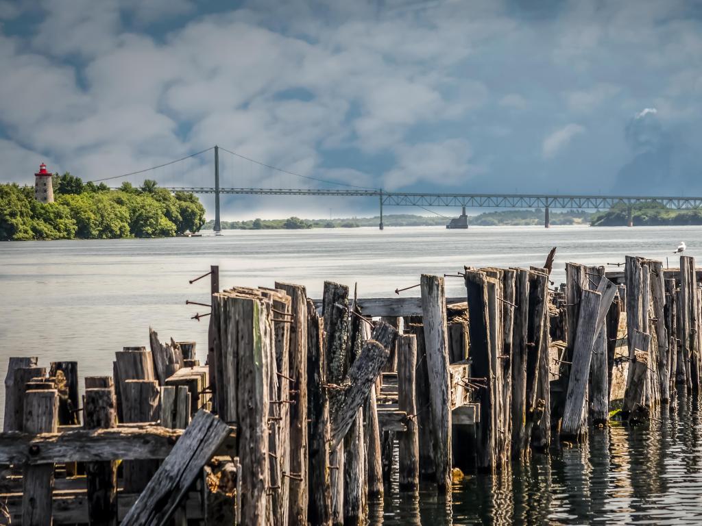 Seaway International Bridge, Canada/USA with antique pilings from an old port with an 1800s lighthouse and the bridge across the St. Lawrence River on a cloudy day.