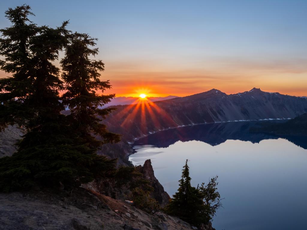 Orange sunset behind the mountains at Crater Lake National Park in Oregon, with pine trees silhouetted in the foreground