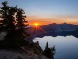 Orange sunset behind the mountains at Crater Lake National Park in Oregon, with pine trees silhouetted in the foreground