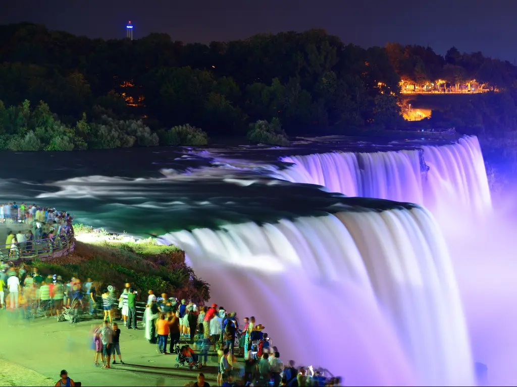 An image where people are looking at the scenic view of Niagara Falls that is enhanced by colorful lights.