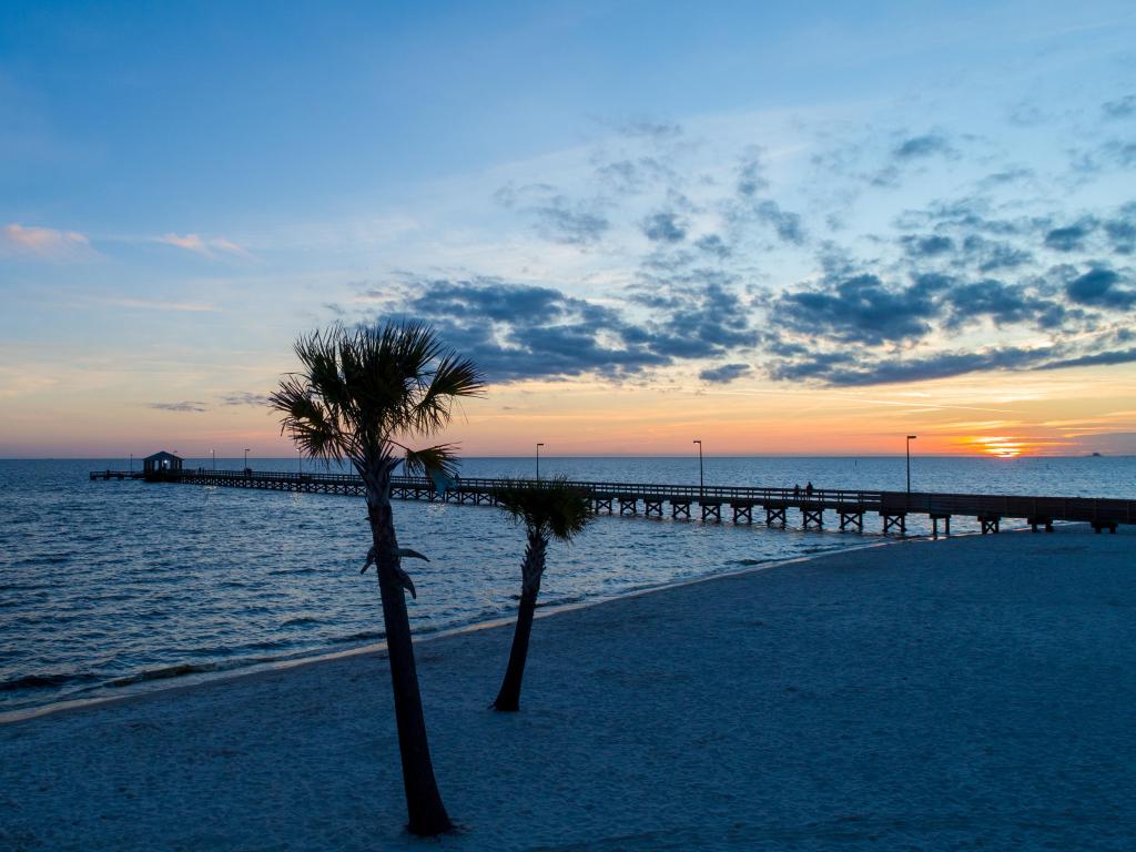 Pier at sunset in Biloxi, Mississippi. Two palm trees are in the foreground, growing in the sand.