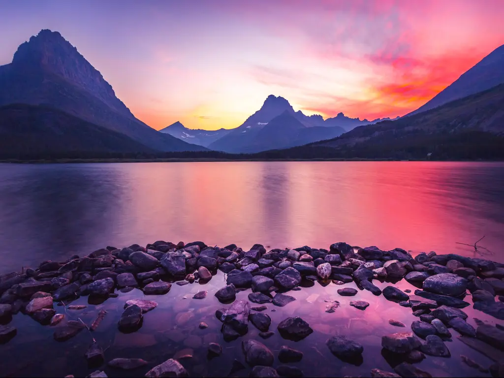 Glacier National Park, Montana, USA at sunset with a dramatic red sky behind mountains in the distance, the calm lake and large rocks in the water in the foreground. 