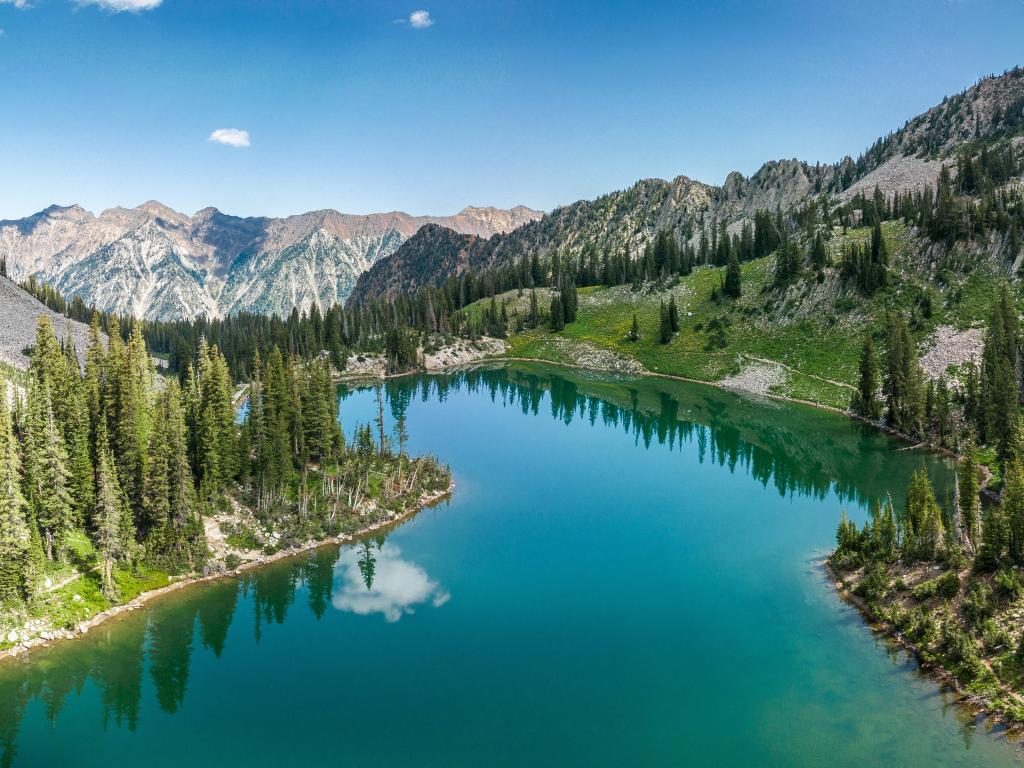 The photo depicts a beautiful alpine lake with blue waters surrounded by tall trees in the Wasatch Mountains on a clear day with only a few clouds in sight.