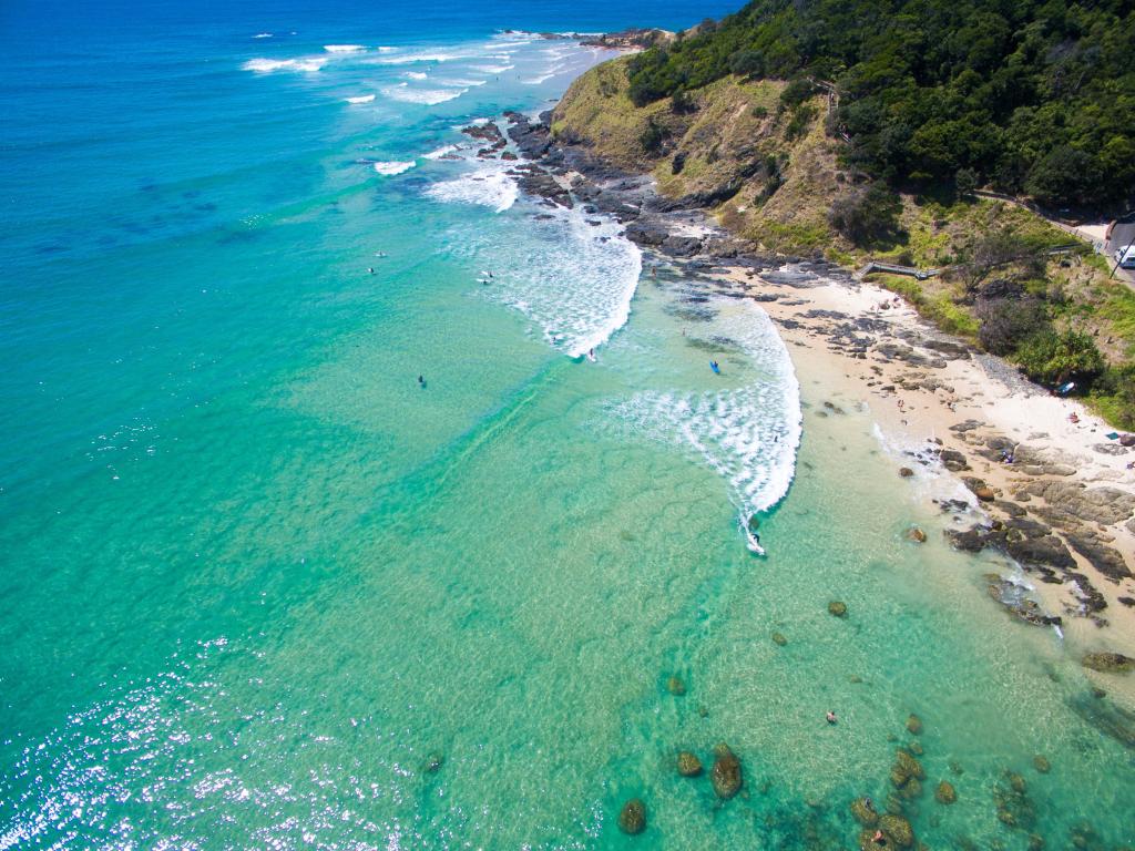 Aerial view of clear blue sea with surfers, with a sandy and rocky beach in the bay and a cliff rising to one side